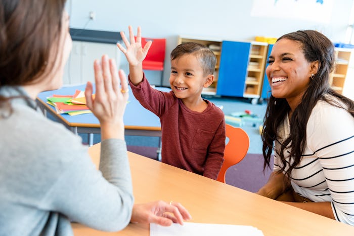 A cheerful teacher gives a little boy a high five during a parent-teacher conference. The boy's mom ...