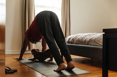 A man in downward dog pose on a yoga mat in his bedroom, with his puppy under him in downward dog.