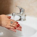 Close-up of a young woman with rings and red nail polish, washing hands in the bathroom sink.