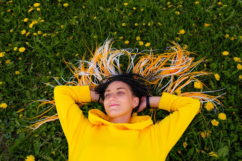 Young woman with long braided yellow hair and blue eyes lying on green grass with dandelions.