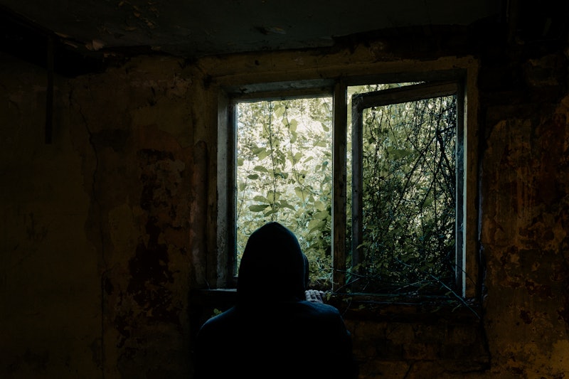 A horror concept. Of a hooded figure, back to camera. Looking out of a broken window. In an abandone...