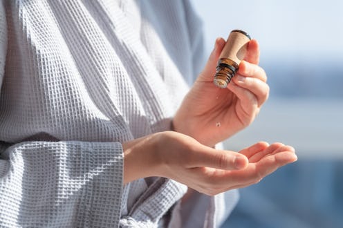 Woman in bathrobe dripping essential oil on hand. Close-up view. Home body care concept. Aroma oil i...