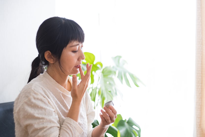 An Asian woman uses CBD cream at home.
She is performing self-care on the window sill of her room.
C...