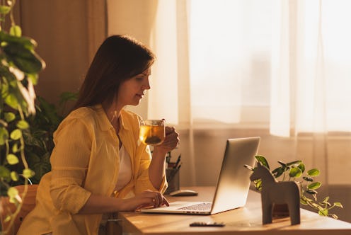 Young business woman planning her day, writing in notepad, sitting in front of laptop, drinking coff...