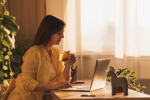 Young business woman planning her day, writing in notepad, sitting in front of laptop, drinking coff...