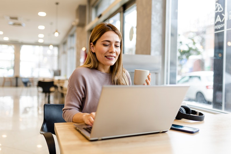 Adult smiling brunette business woman in stylish shirt working on laptop in cafe
