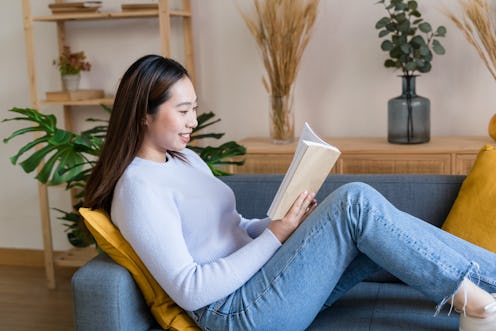 Side view of young female in casual clothes sitting on sofa and reading book