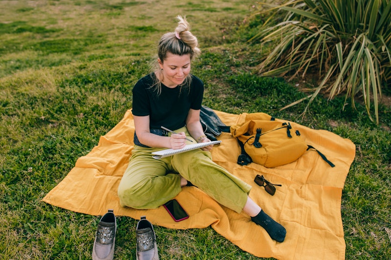 Young beautiful woman drawing on a sketchbook sitting on a blanket in a public park