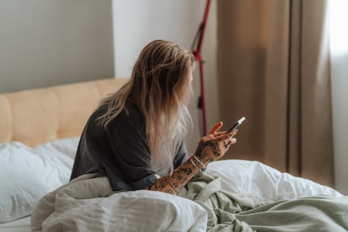 Close up blond woman with messy hair struggles to wake up while surfing the net on her smartphone in...
