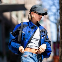 Hailey Bieber wears a gray t-shirt with baggy jeans and a blue motocross jacket and baseball cap