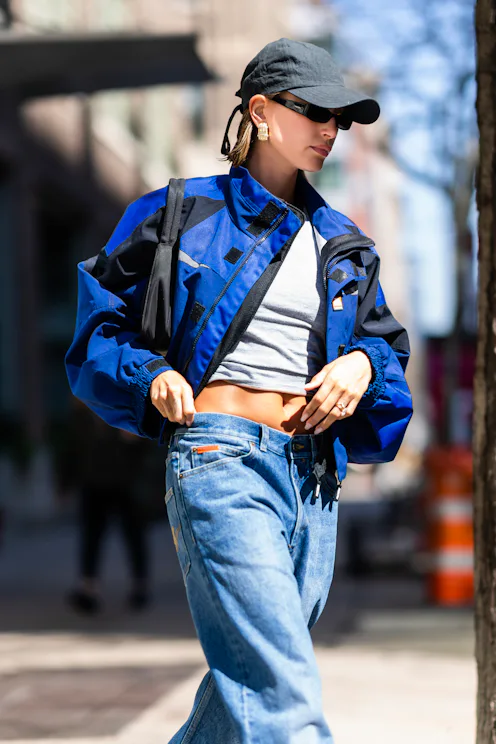 Hailey Bieber wears a gray t-shirt with baggy jeans and a blue motocross jacket and baseball cap