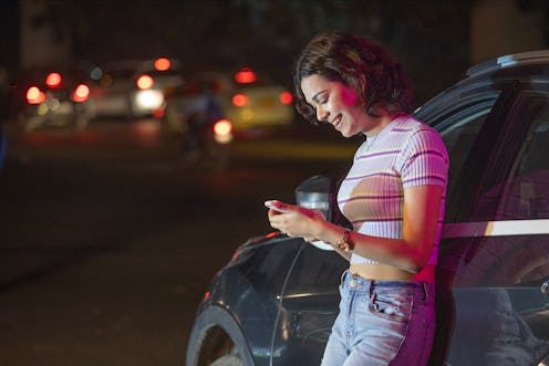Smiling young woman leaning on car and text messaging using mobile phone outdoors at night