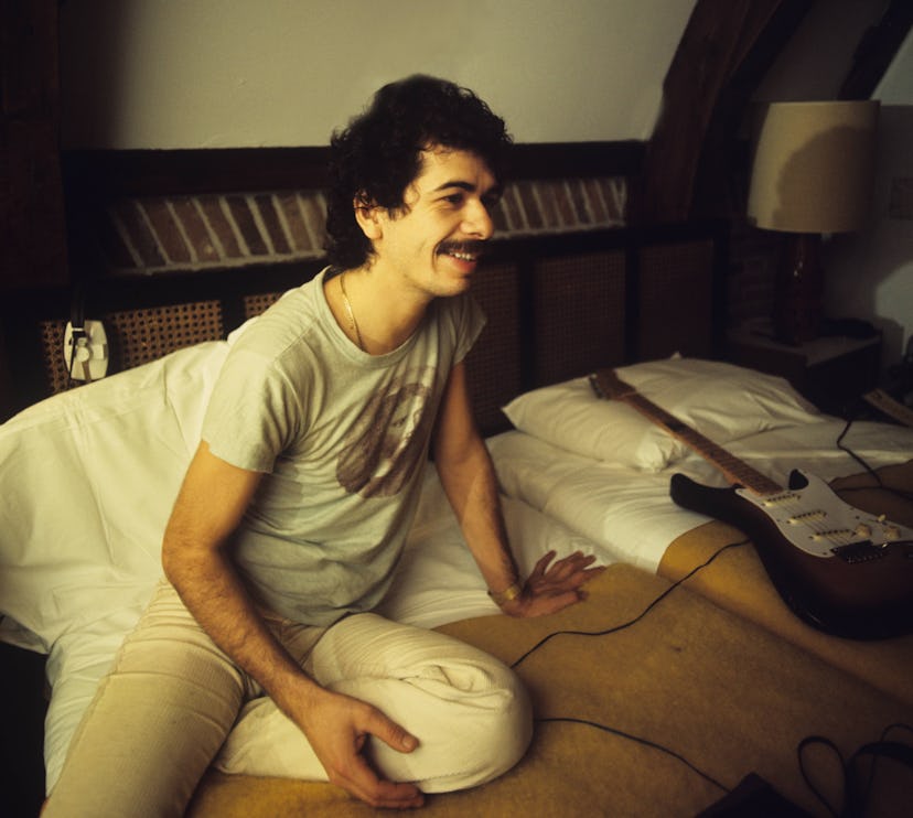 A portrait of Carlos Santana, inspiration for many baby names, on a hotel bed with a Fender Stratoca...