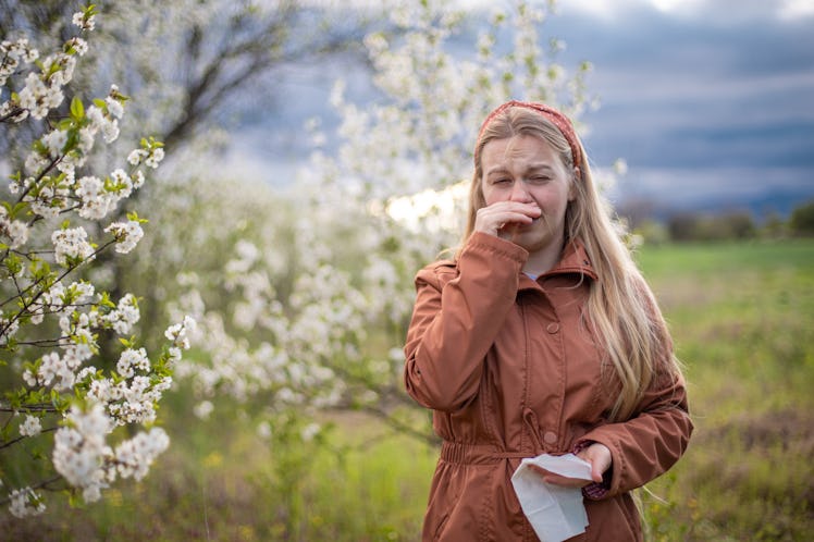 Young woman walking next to blooming tree and having allergic reaction of pollen. She is holding tis...