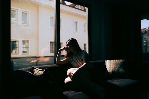 A tired and depressed Mediterranean woman is sitting on the sofa near the window at home, feeling ex...