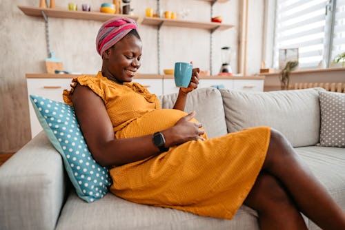 Beautiful pregnant black mother holding her pregnant belly at home and drinking tea on sofa in artic...