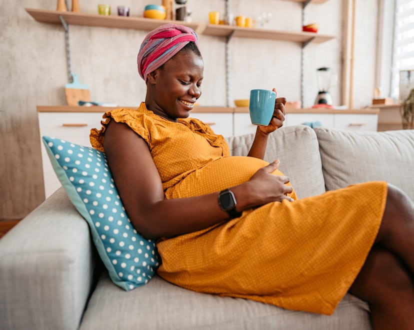 Beautiful pregnant black mother holding her pregnant belly at home and drinking tea on sofa in artic...