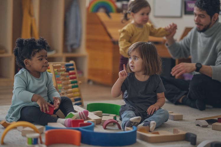 Daycare children are seen sitting on the floor as they pay with wooden blocks together. The toddler...