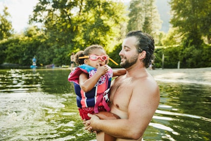 Father holds young daughter, wearing life jacket and goggles, in a lake, in a story about how to cho...