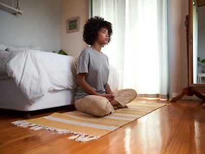 Young African woman sitting with her eyes closed and meditating on her bedroom floor during a yoga s...