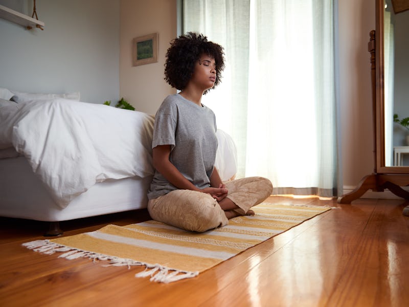 Young African woman sitting with her eyes closed and meditating on her bedroom floor during a yoga s...