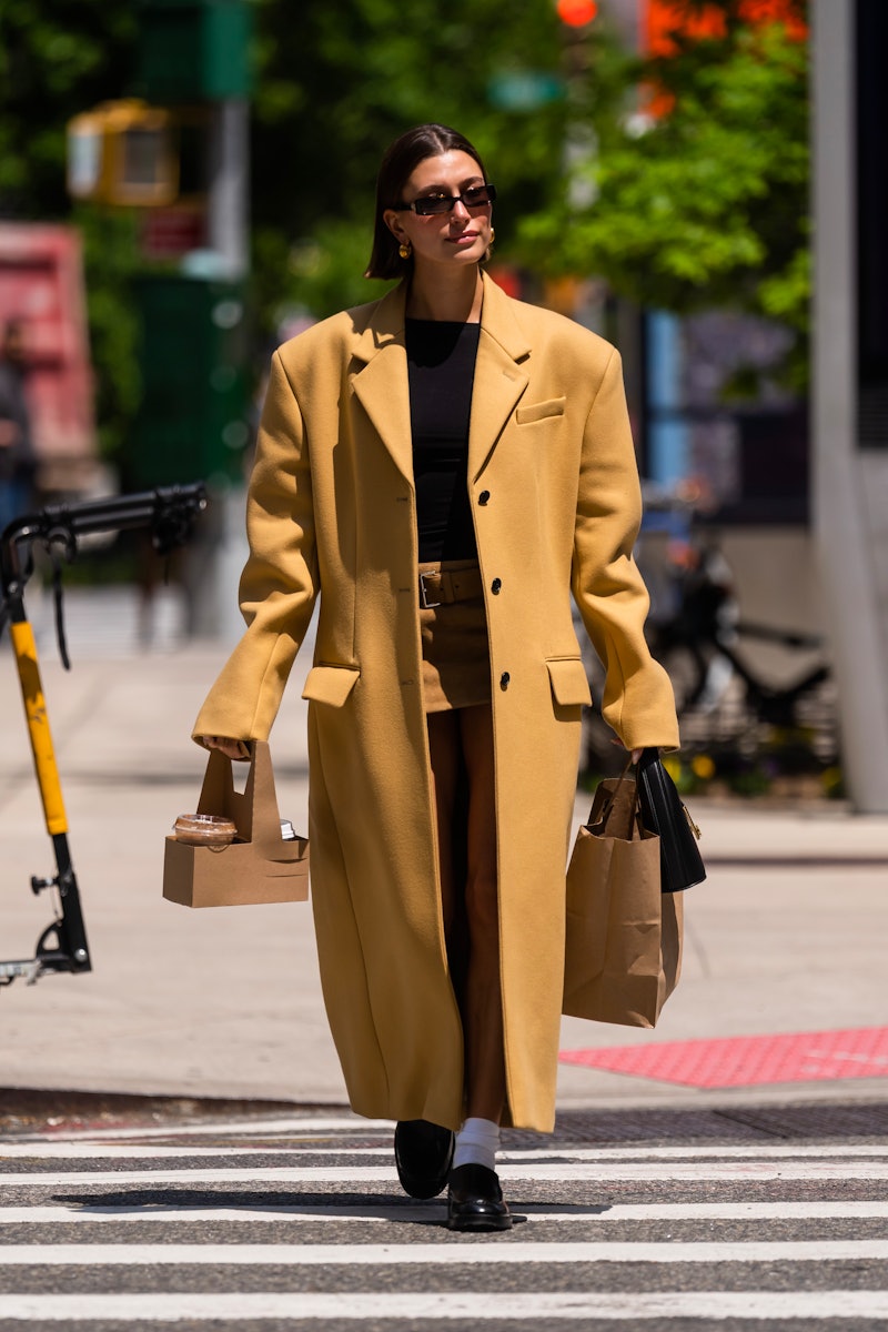 Hailey Bieber wears a camel top coat with a micro mini khaki skirt and black top with loafers to get...