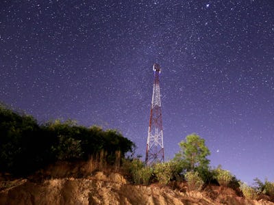 A red and white cell phone tower on a tree-lined cliff against a starry night sky