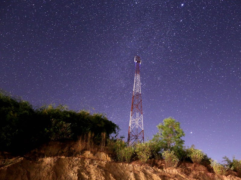 A red and white cell phone tower on a tree-lined cliff against a starry night sky