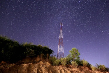 A red and white cell phone tower on a tree-lined cliff against a starry night sky