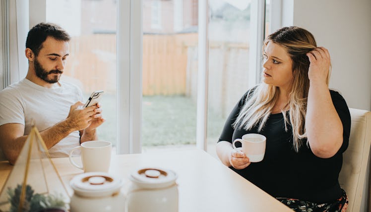 A woman and man sit at a kitchen table. The man looks down at his phone and subsequently ignores the...