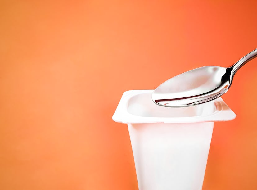 Yogurt cup and silver spoon on orange background, white plastic container with yoghurt cream, fresh ...
