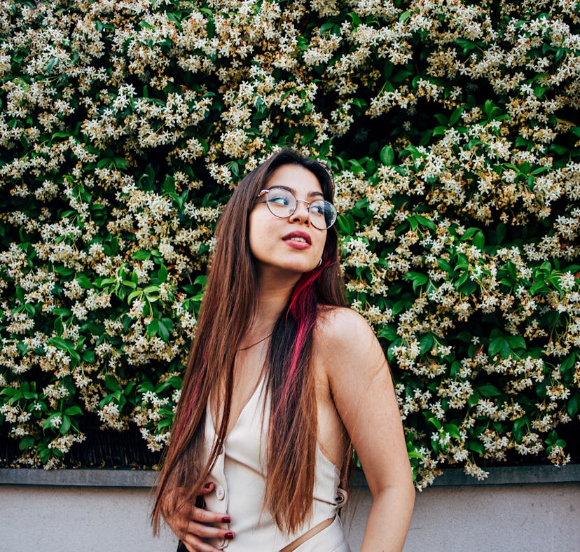 young woman with pink highlights poses in front of a large flower bush as she considers the spiritua...