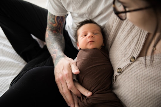 Goth baby names would fit this newborn baby, who sleeps in a swaddle in Mom's arms while Dad looks o...