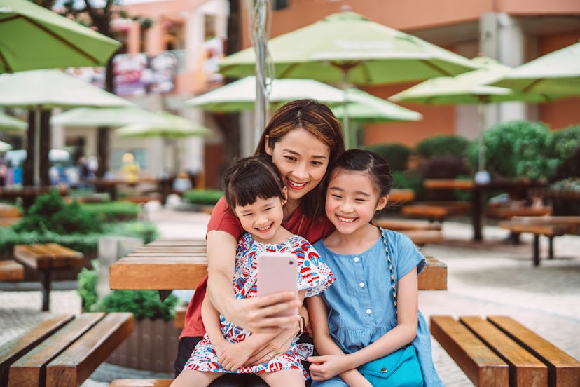 Pretty young mom taking selfie joyfully with her two lovely daughters using smartphone while having ...