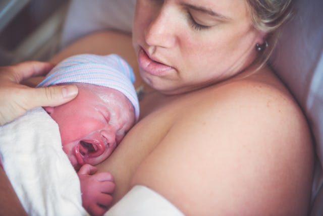A young mother lays in her hospital bed with her crying newborn baby on her chest.