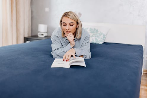 A young Caucasian woman is lying on her stomach, enjoying a book on her bed.