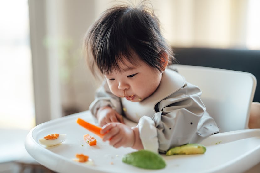 When can babies eat eggs? Baby girl eats a steamed carrot off her high chair tray, with eggs and avo...