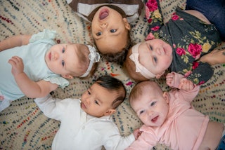 Five babies lay on the floor in a circle with their heads close together as they pose for a portrait...