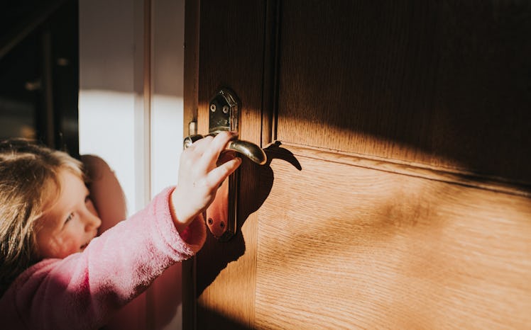 Cute little girl reaches up to open a door handle that is too high for her. She smiles knowingly at ...
