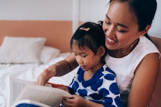 Asian mother and her little girl sitting comfortably on the bed at their home, focused on reading a ...