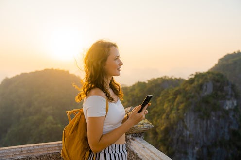 Young Caucasian woman standing on the top of wat Tham Sua in Krabi at sunset and using smartphone