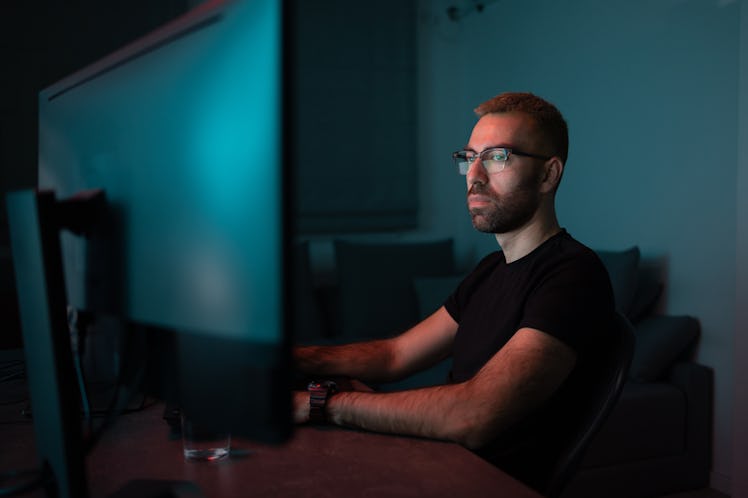 Side view of a man in his thirties with dyed hair, a nose ring, and glasses working on a computer at...