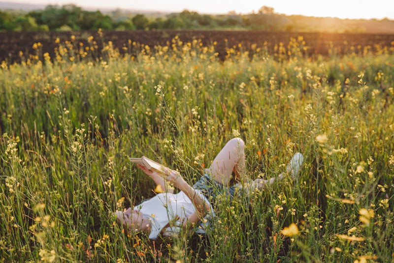 Young woman lies on a flowering meadow in the grass and reading a book