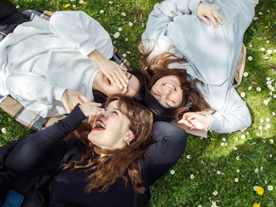 Group of young women with brown wavy hair in comfortable sweatshirts enjoying outdoors, lying on gra...