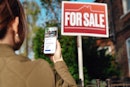 Rear view of woman looking using smartphone while looking at real estate sign, planning to buy a hou...