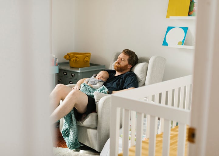 A dad and an infant have fallen asleep together sitting in a chair in the baby's nursery; you can se...