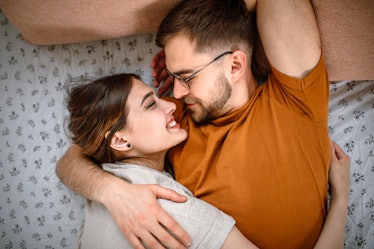 A young heterosexual couple embrace in bed, the man wears an orange t-shirt and glasses and the woma...