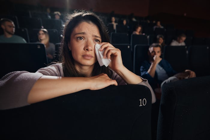 Depressed woman wiping her tears while watching a sad movie in theatre. There are people in the back...