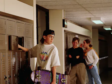 A teenager in the '90s stands at his locker: he's wearing a backwards hat, a vintage tee, a flannel ...