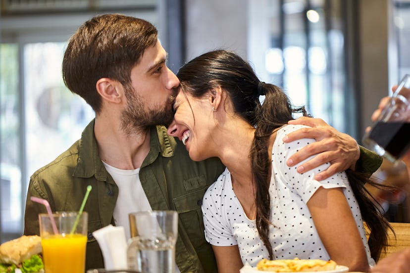 Front view of young adult man sitting at mall's food court table while kissing girlfriend's fronthea...